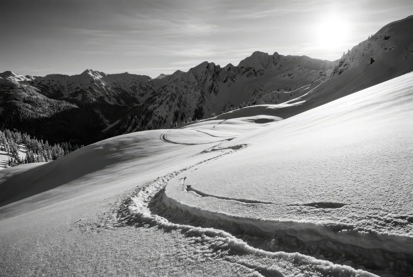 A single ski track running down toward a low sun over distant ridges